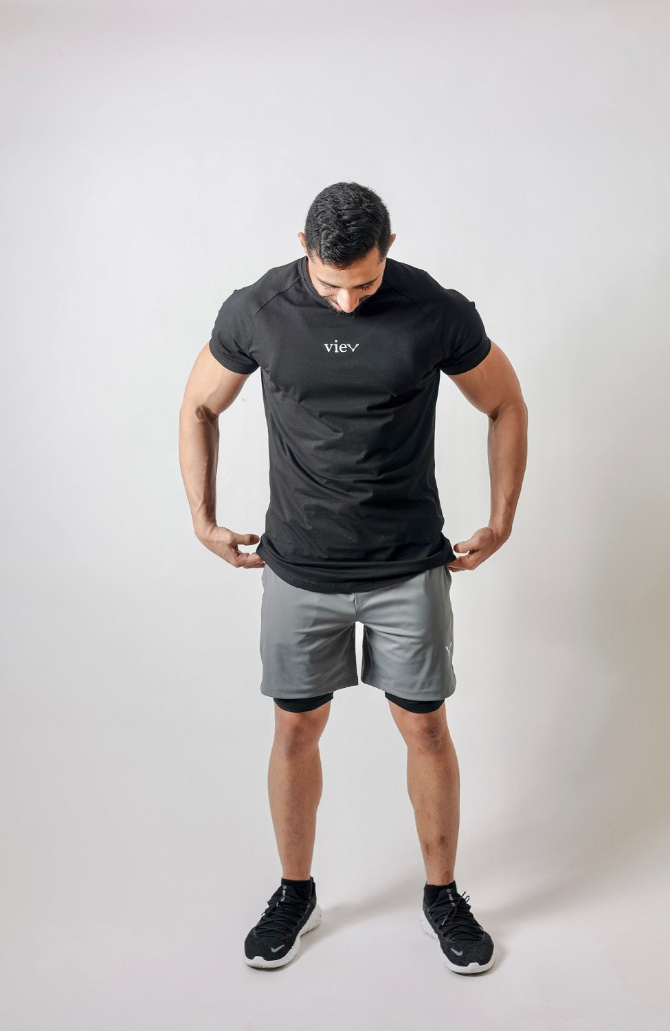 Model looking down while holding the hem of a black Vie T-shirt worn with grey shorts.